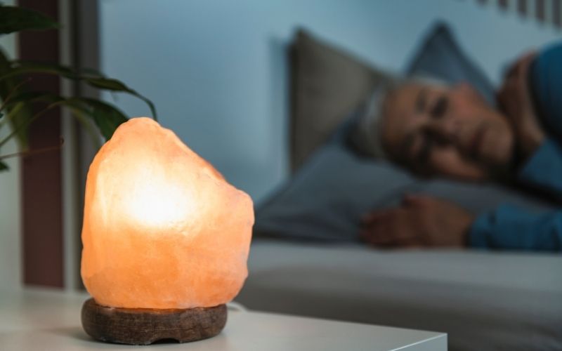 a salt lamp at bed side with a sleeping woman on a bed.